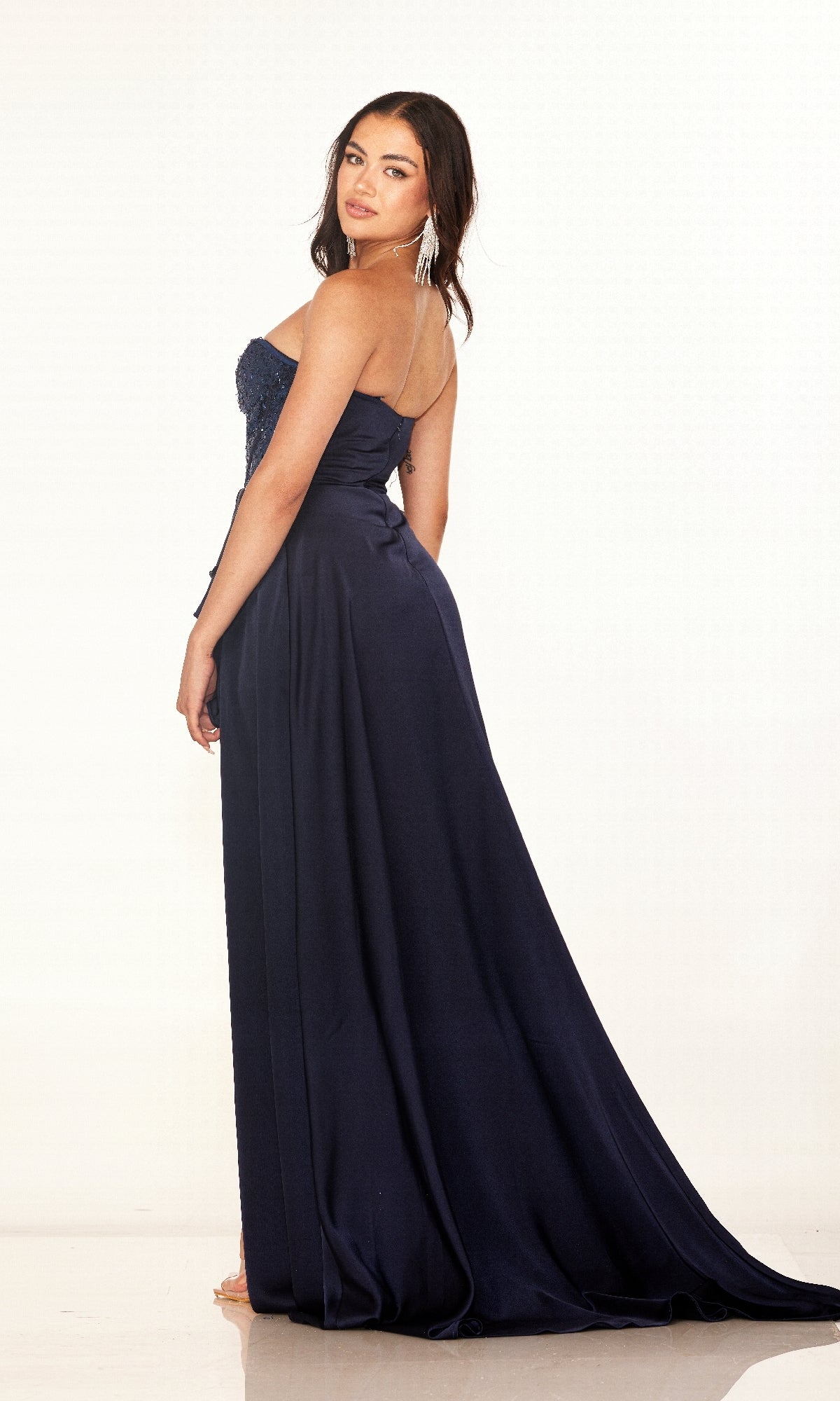 A woman models the Long Prom Dress: Cecilia Couture 2607, featuring a strapless sequin corset bodice and floor-length navy skirt, as she poses against a plain white background, glancing over her shoulder.